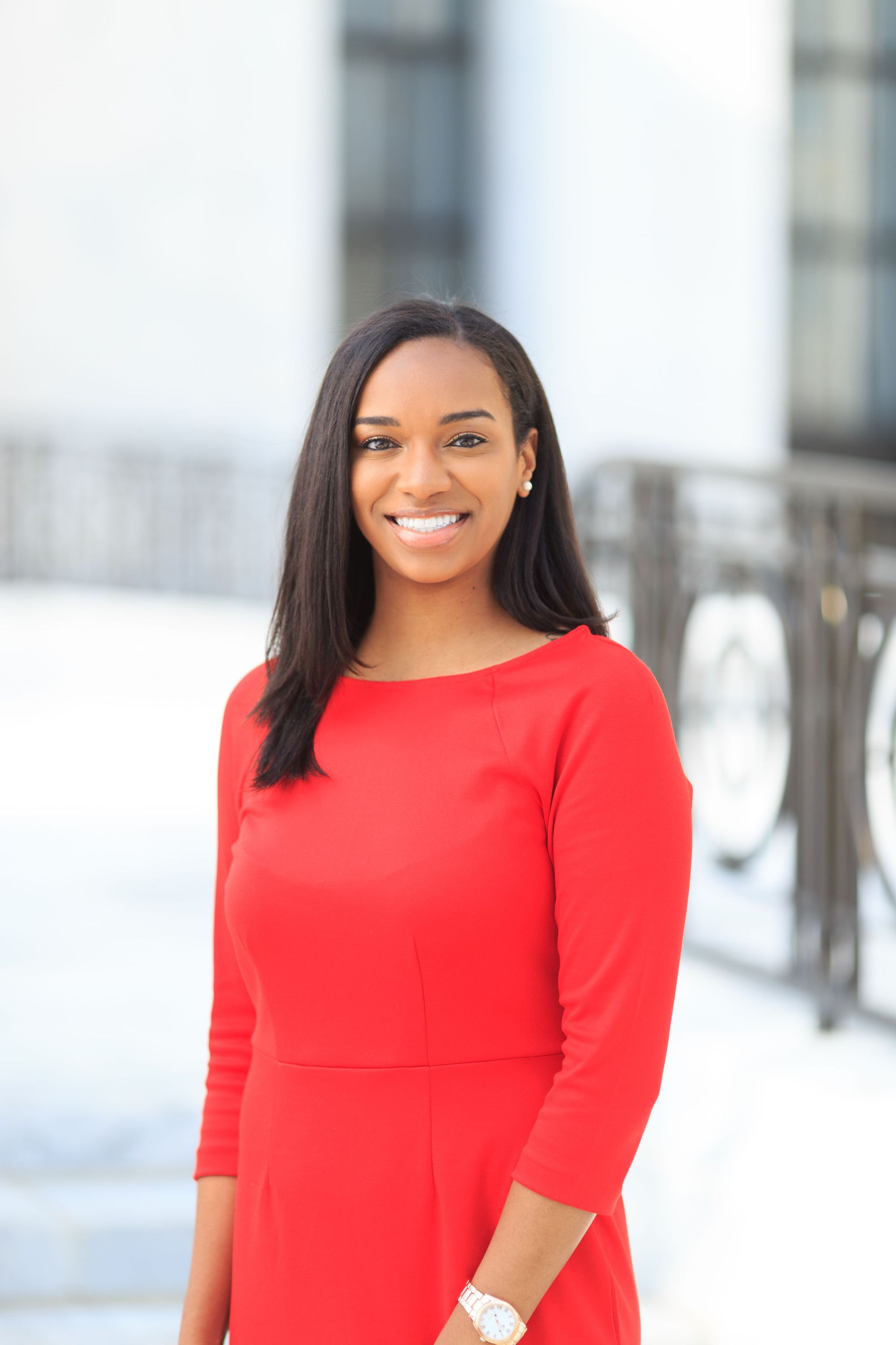 A smiling person with long dark hair wears a bright red, three-quarter sleeve dress, standing outdoors.