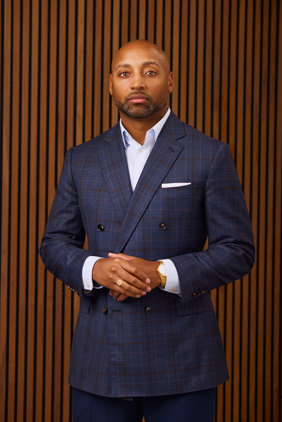 A person in a blue plaid double-breasted suit and light shirt, standing with hands clasped against a wood-slat background.