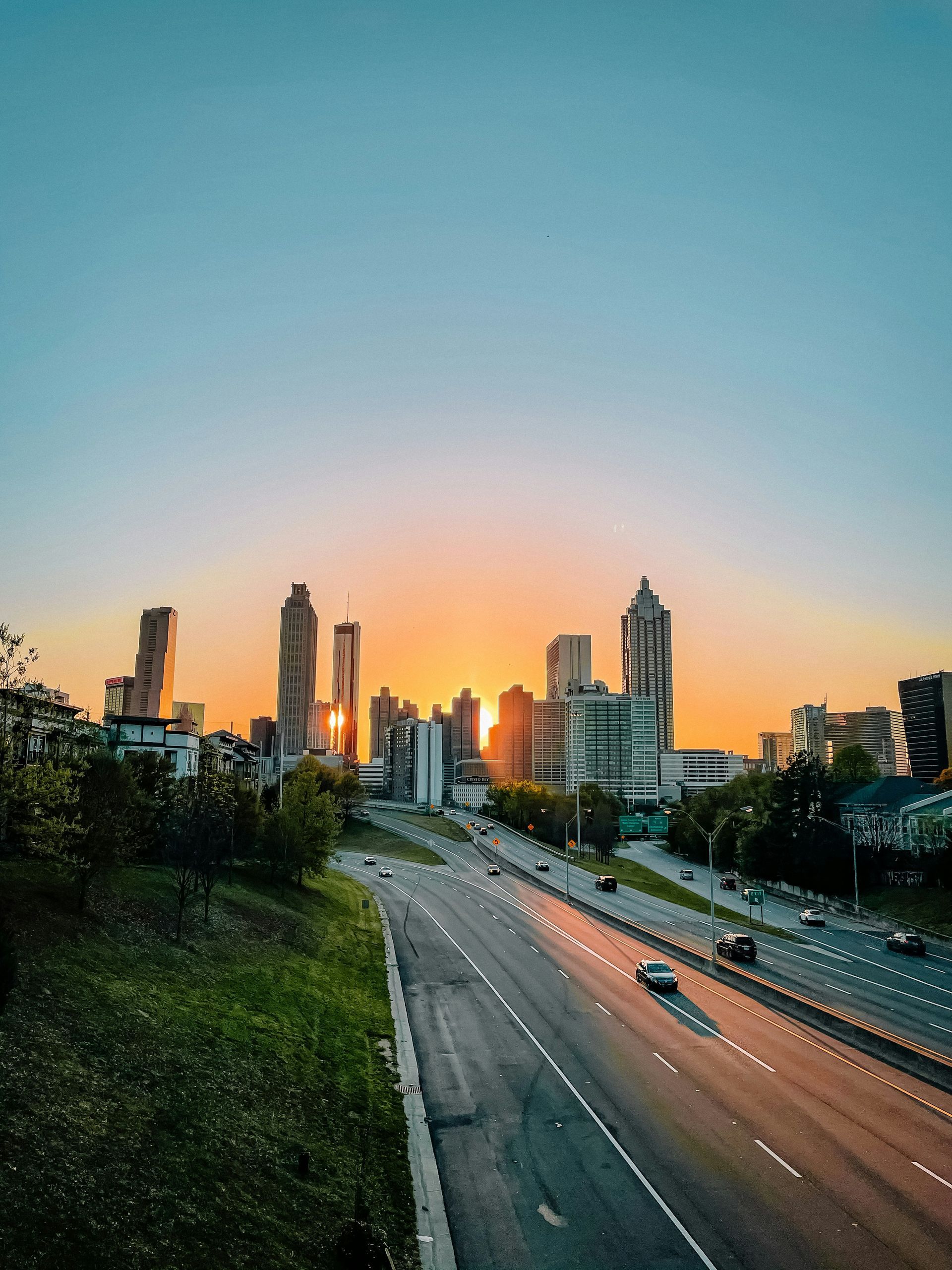 A highway leads toward the illuminated Atlanta skyline at sunset, with cars driving under a gradient blue and orange sky.