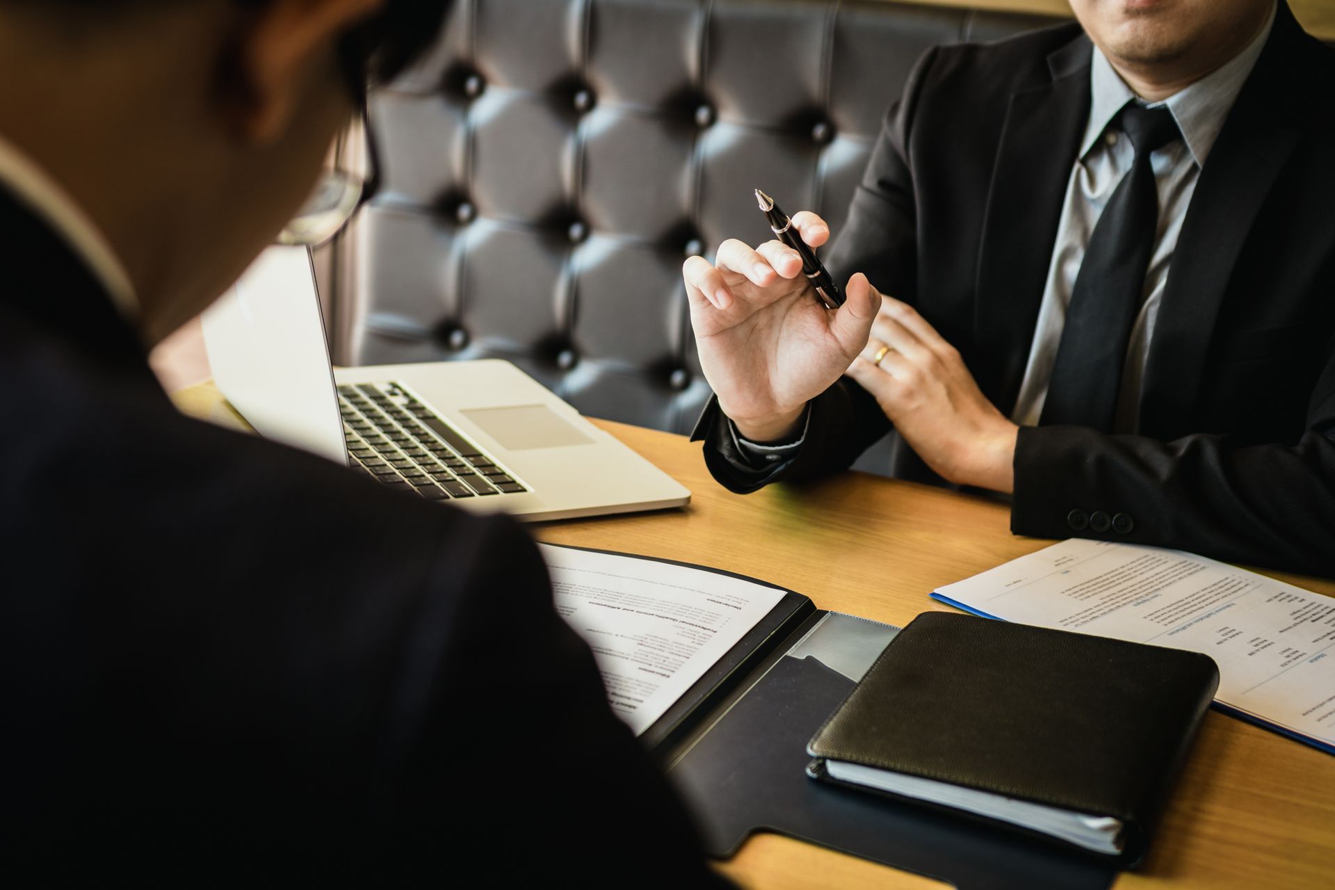 Two professionals in suits discuss business with documents and a laptop on a wooden desk in a modern office.