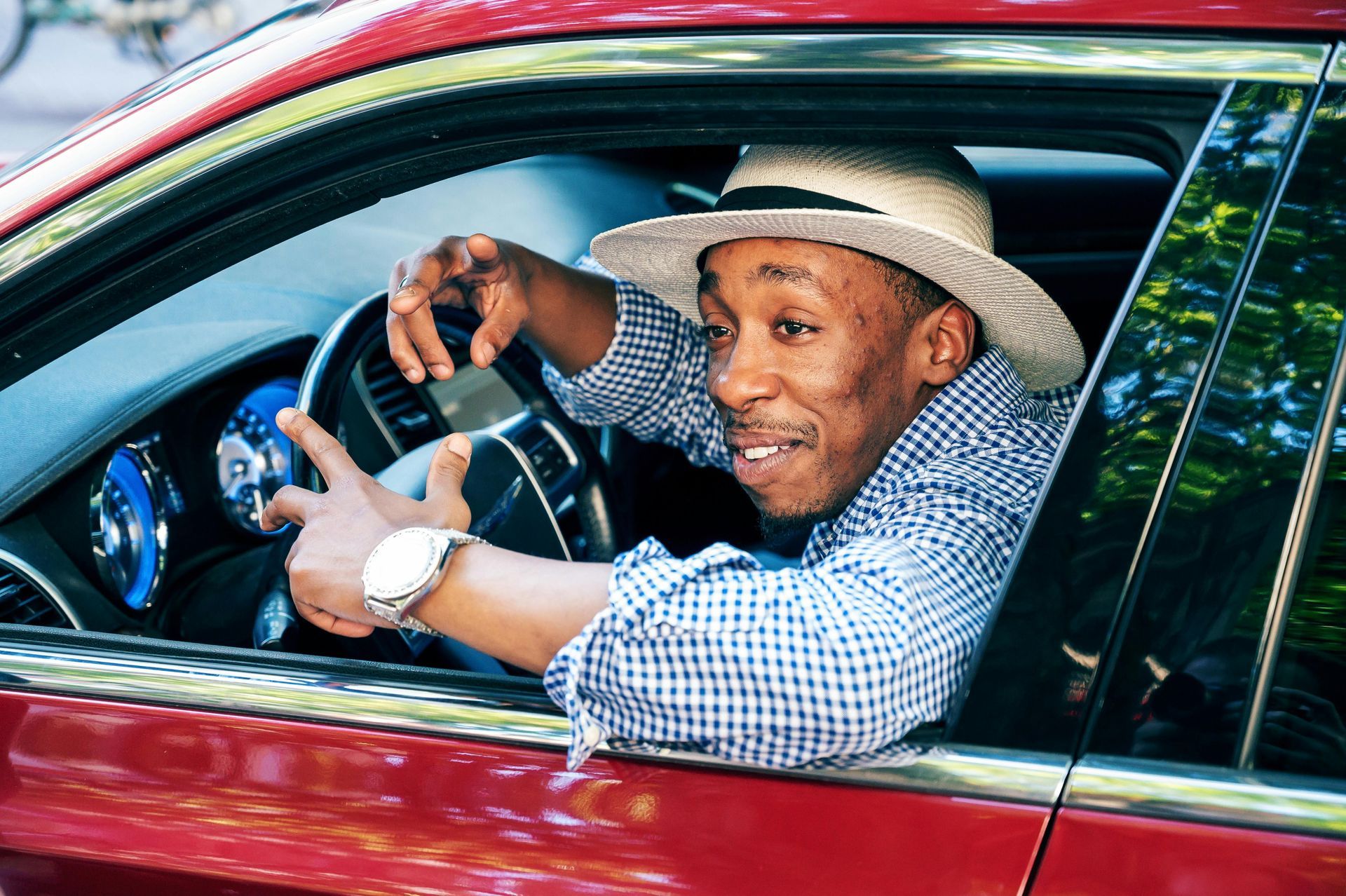 A person in a straw hat and blue gingham shirt sits in the driver's seat of a red car, gesturing while smiling.