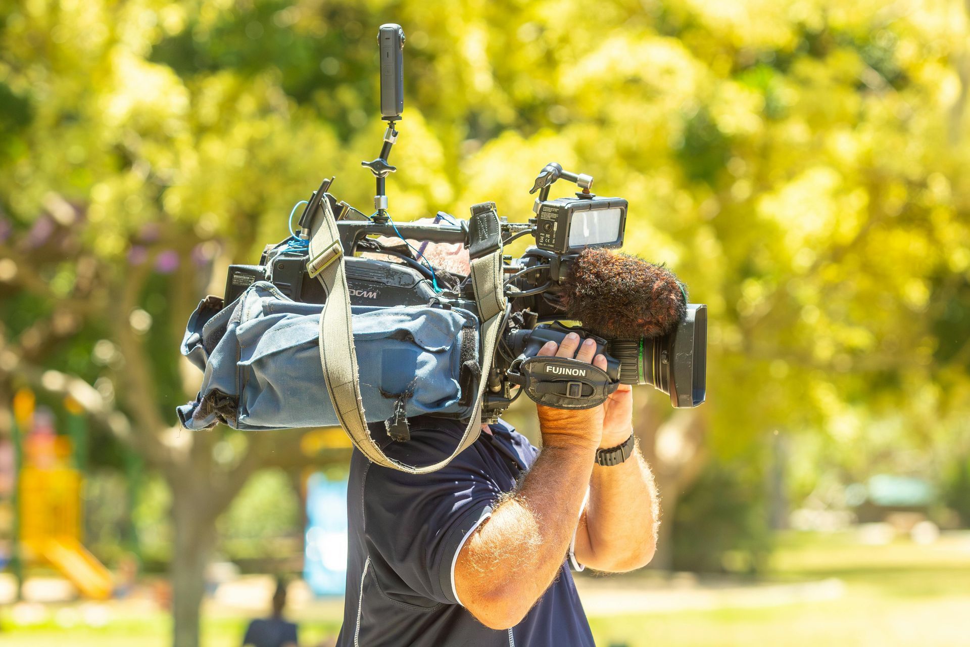 A camera operator holds a professional shoulder-mounted video camera while filming outdoors in a sunlit park.