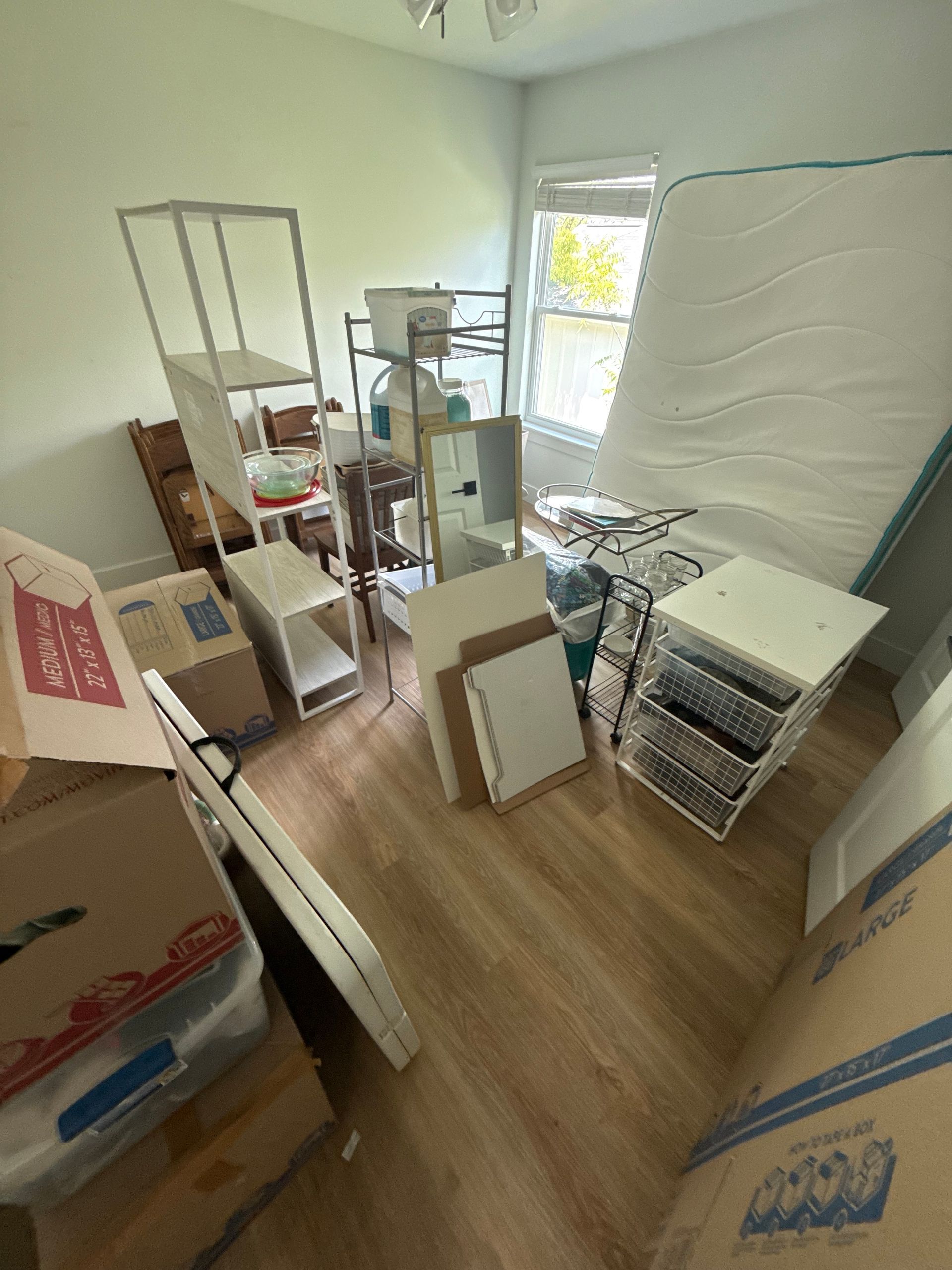 Room cluttered with furniture and boxes, likely during a move; items include shelves, mattress, mirror, and boxes.