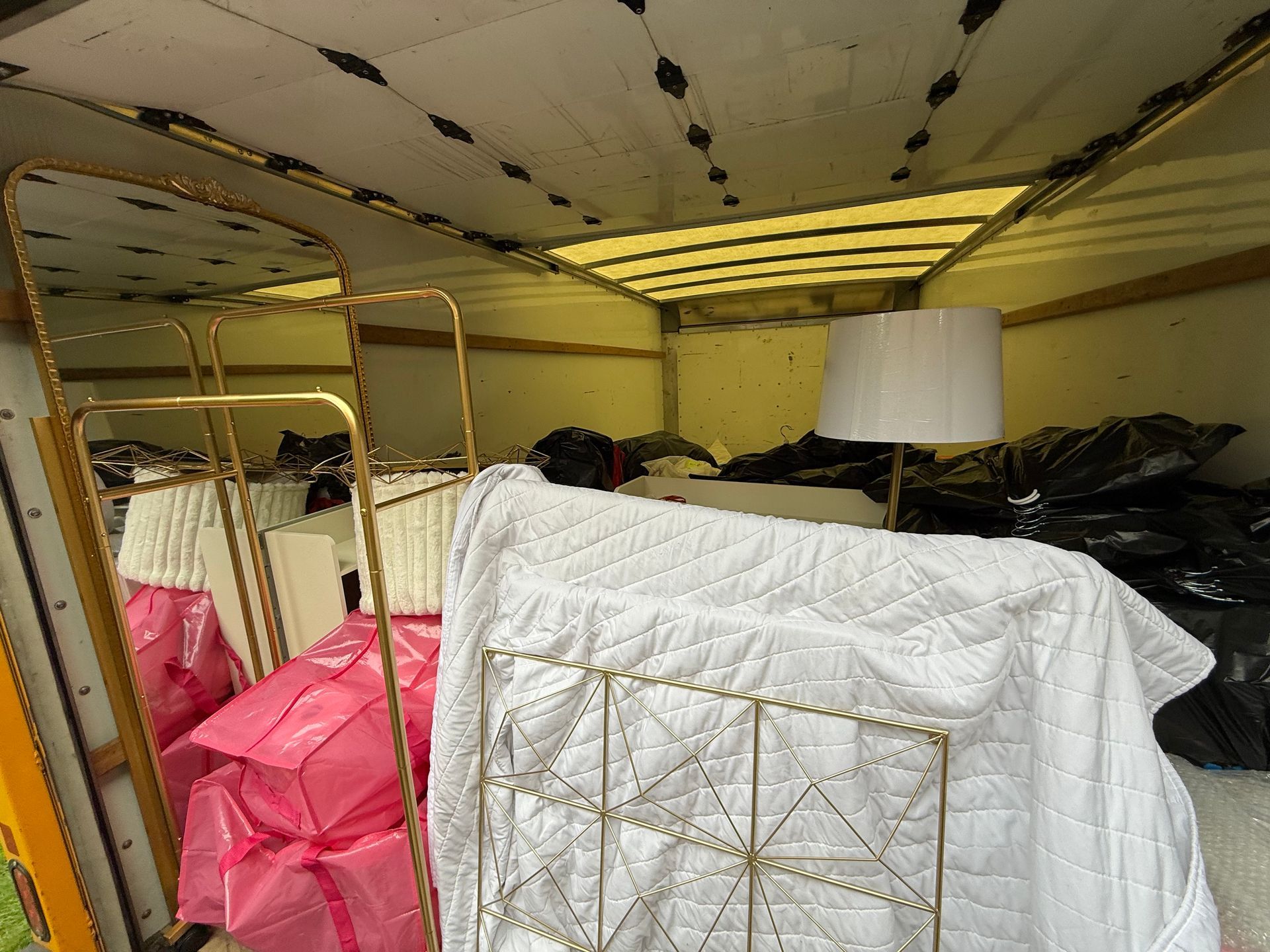 Inside a moving truck, a gold-framed mirror, bed frame, and lamp are among the packed furniture.