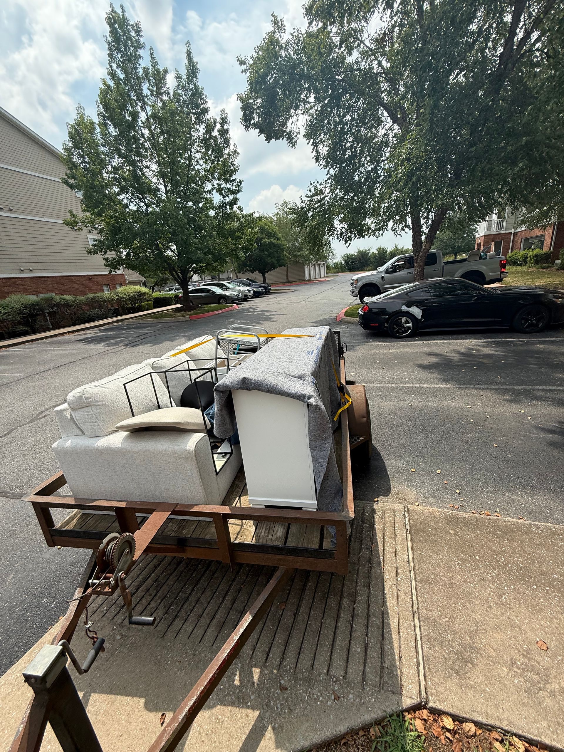 A trailer loaded with furniture parked on a paved driveway near apartment buildings.