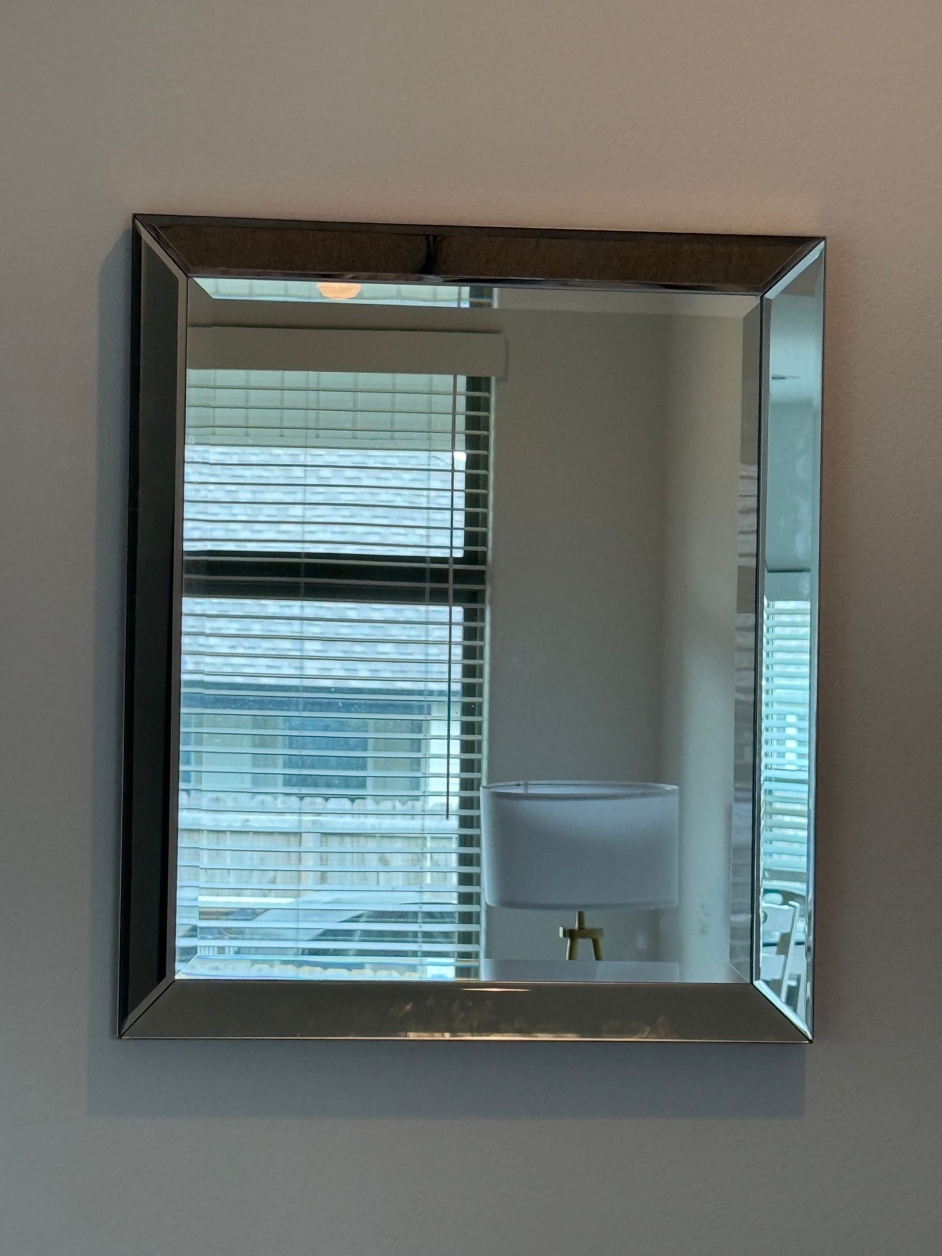 Square mirror with silver frame reflects a window with blinds, and a lamp.