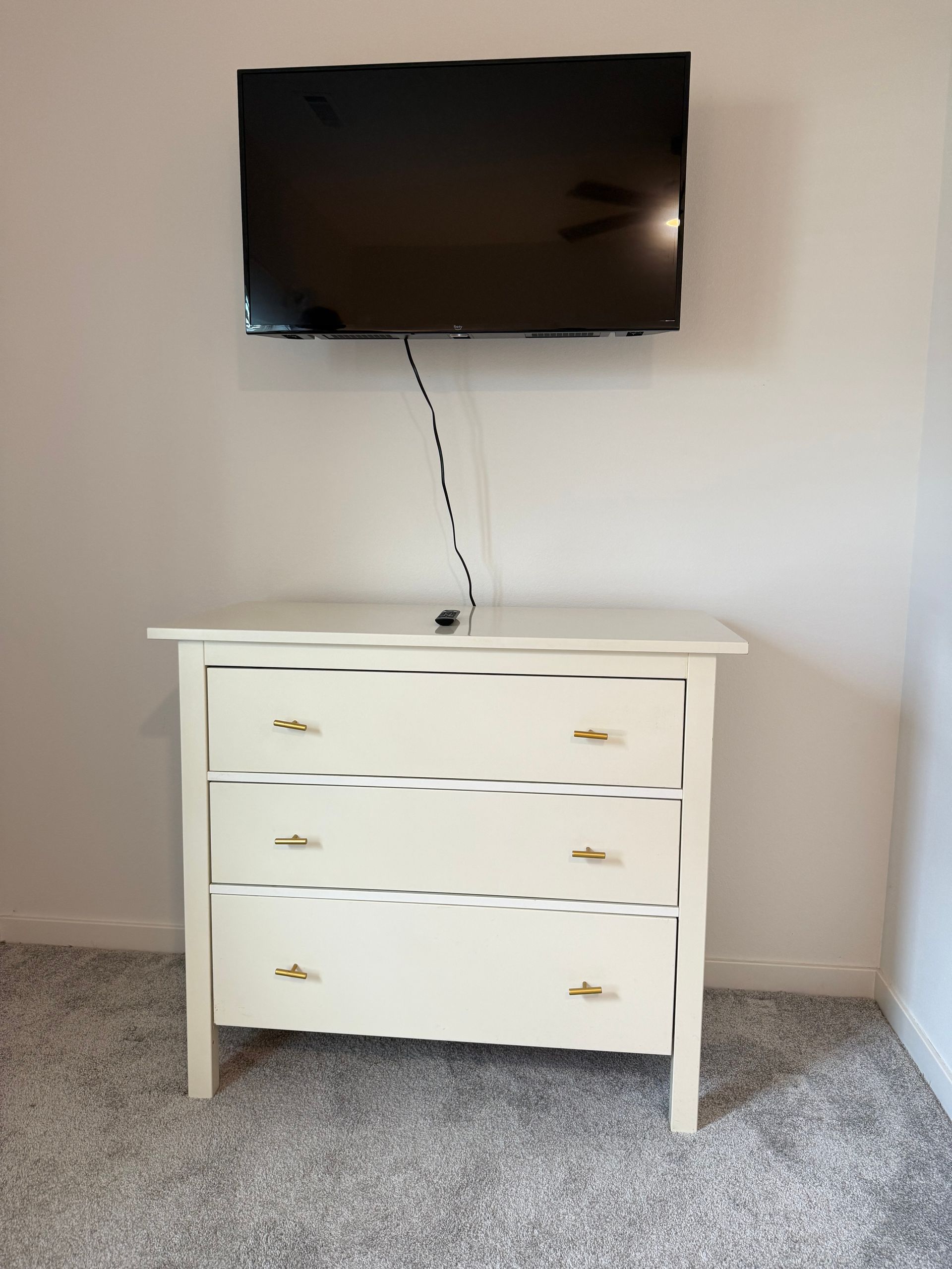 White dresser with gold handles under a wall-mounted TV. Gray carpet and white walls.