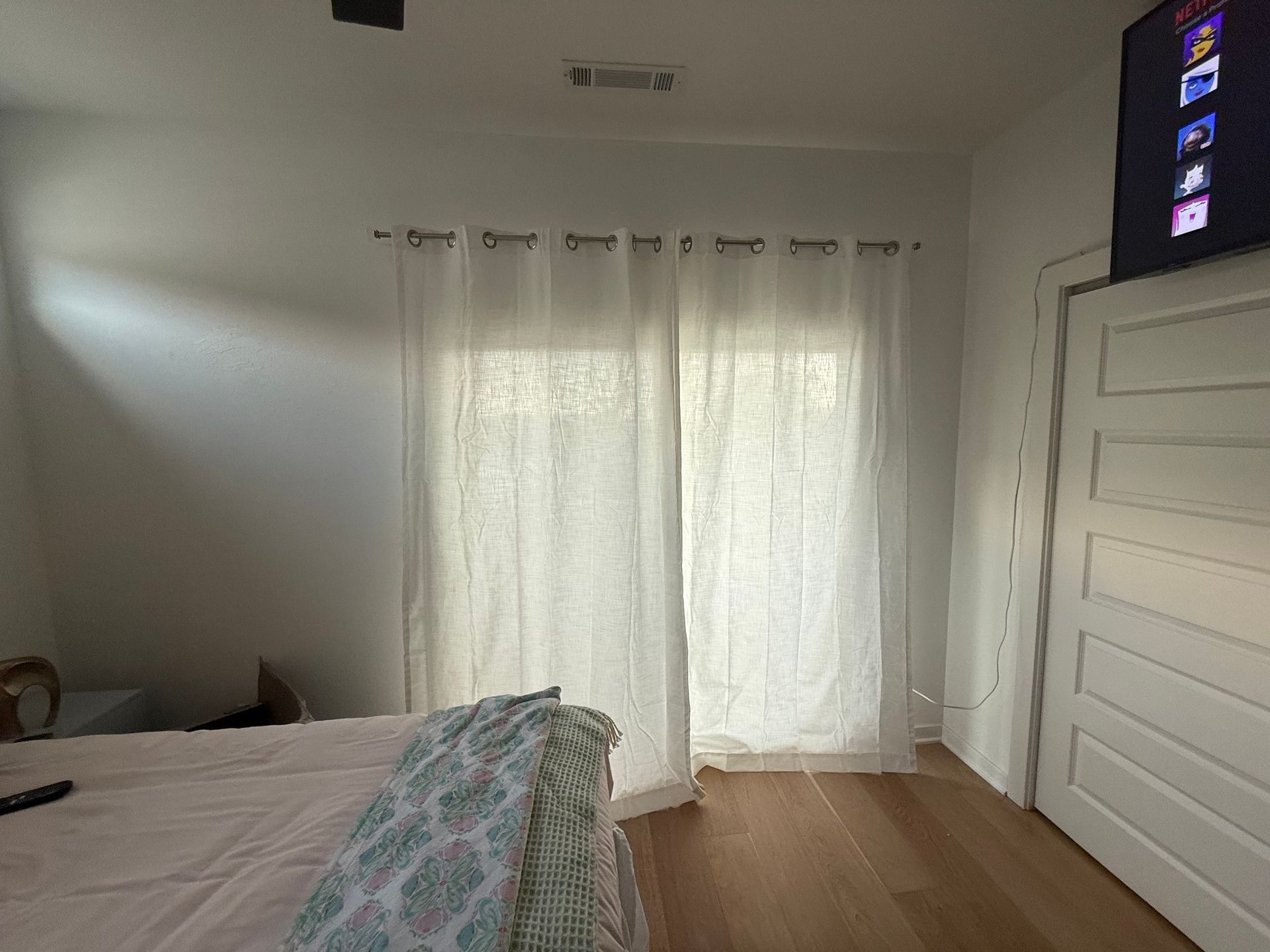 Bedroom with white sheer curtains over a sliding glass door, bed with a patterned throw, and a TV.