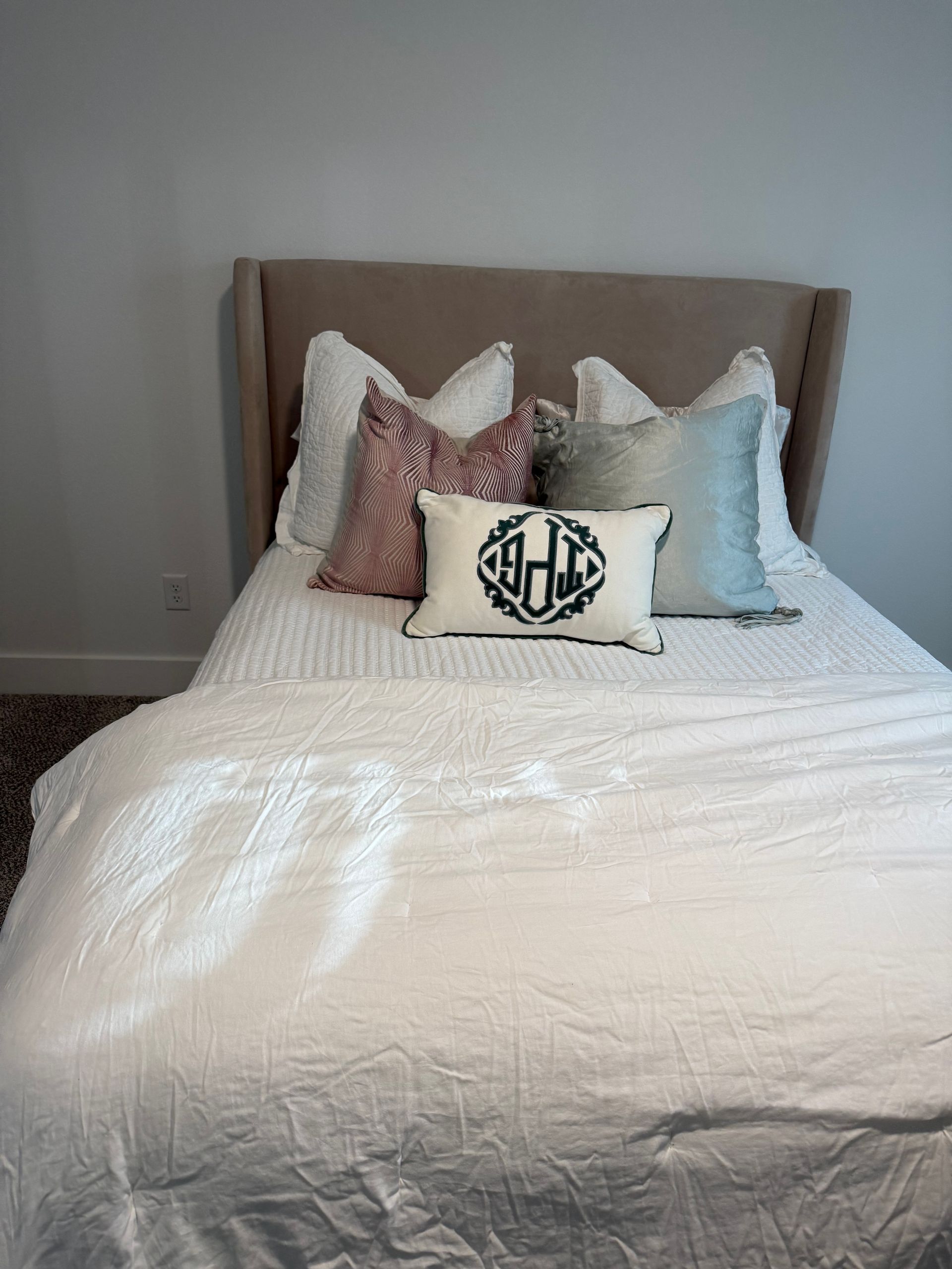 Bed with light-colored linens, pillows, and headboard. One pillow has a monogram design.