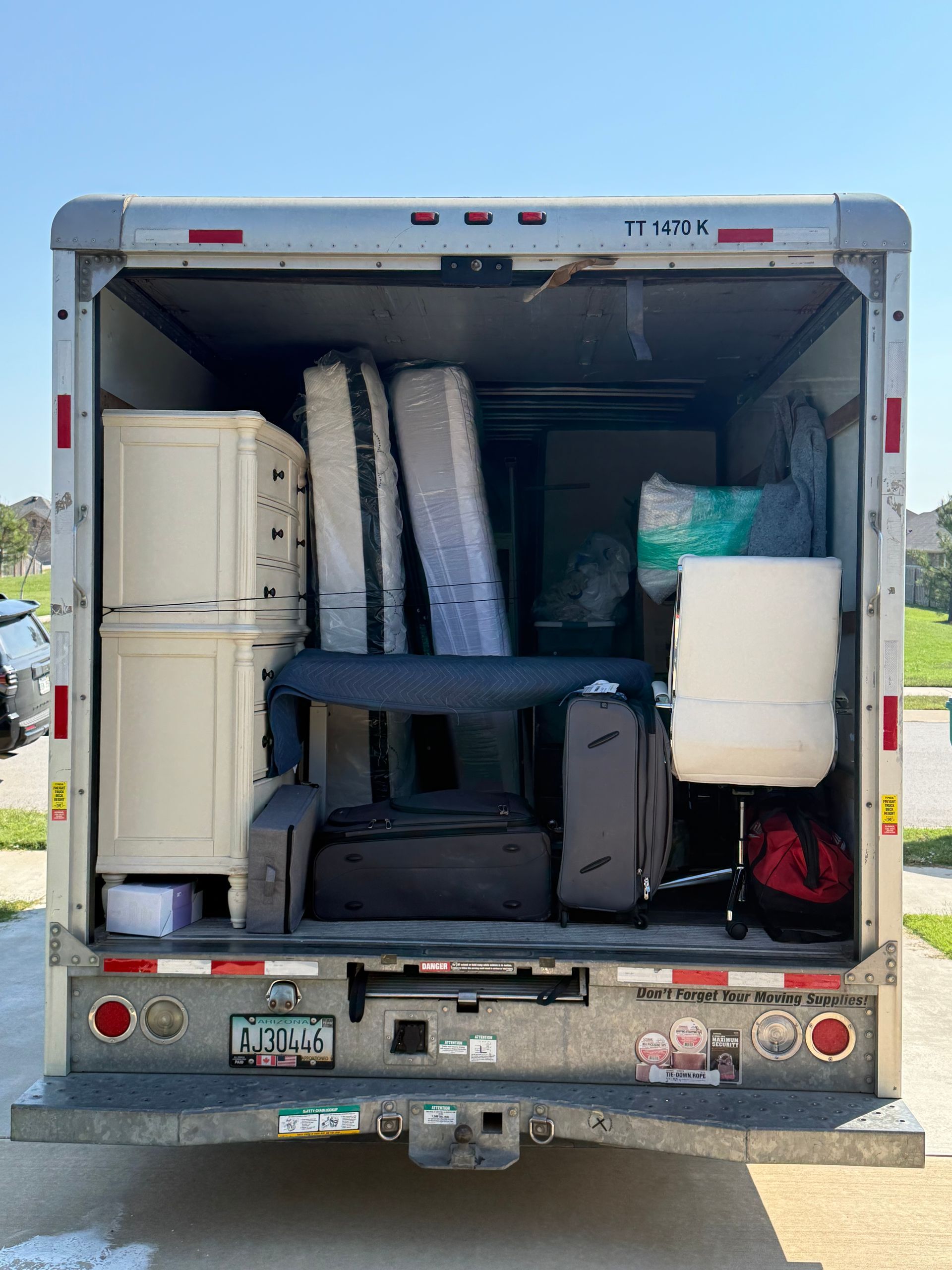 Rear view of a moving truck loaded with furniture, including cabinets, mattresses, luggage, and a desk.