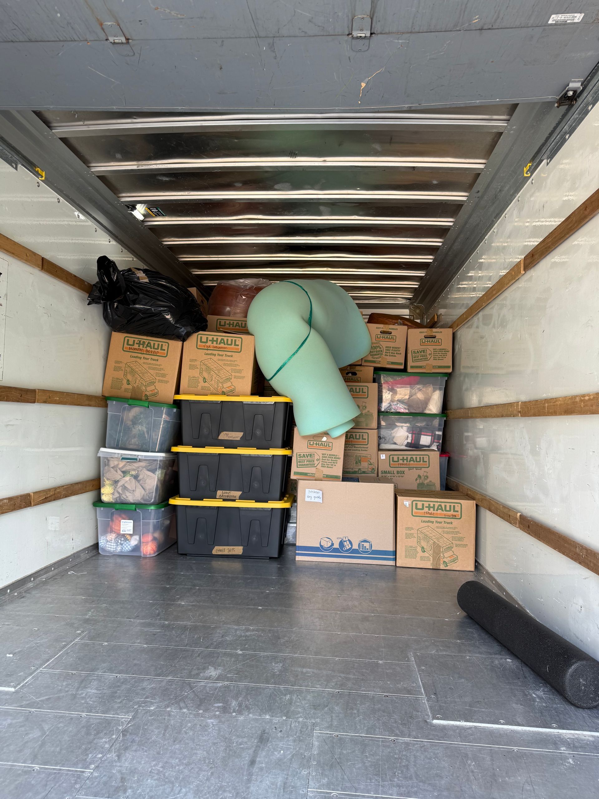 Inside of a moving truck filled with cardboard boxes, bins, and a rolled rug.