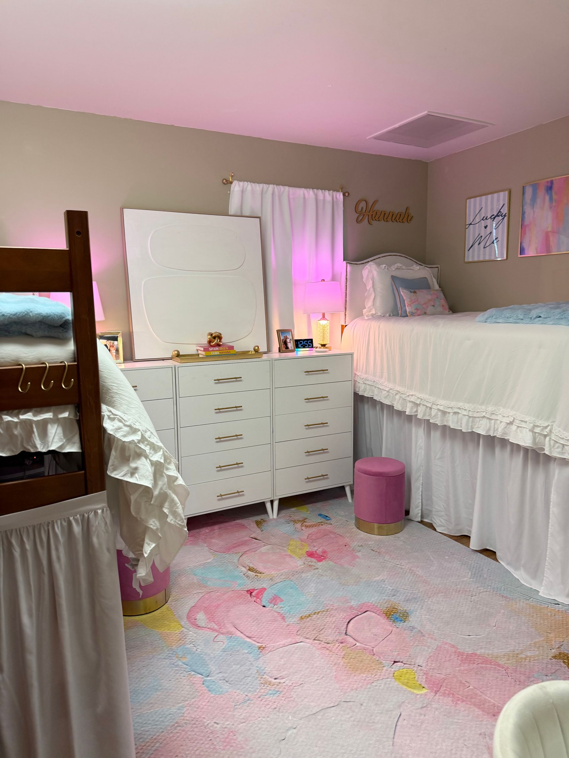 A dorm room with white furniture, a colorful rug, and pink lighting.