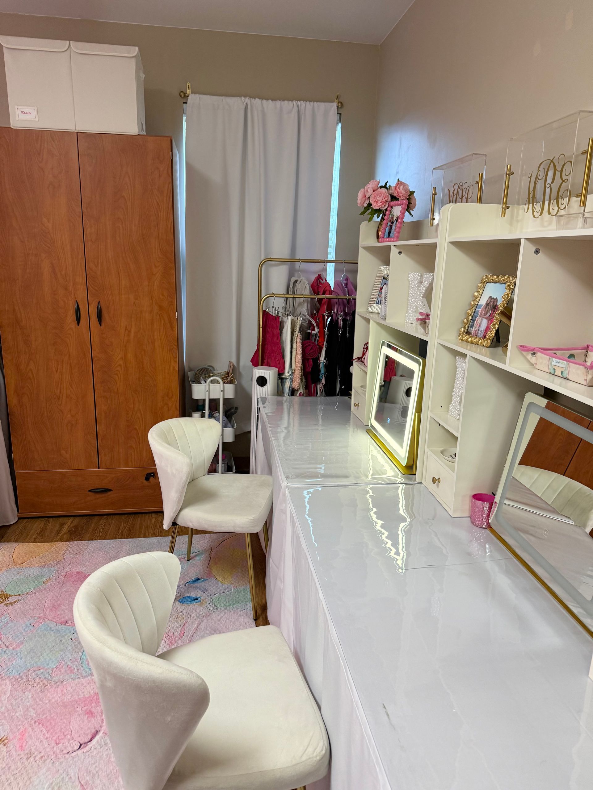 Vanity room with white desk, chairs, and wardrobe; pink clothing rack, flowers, and shelves.