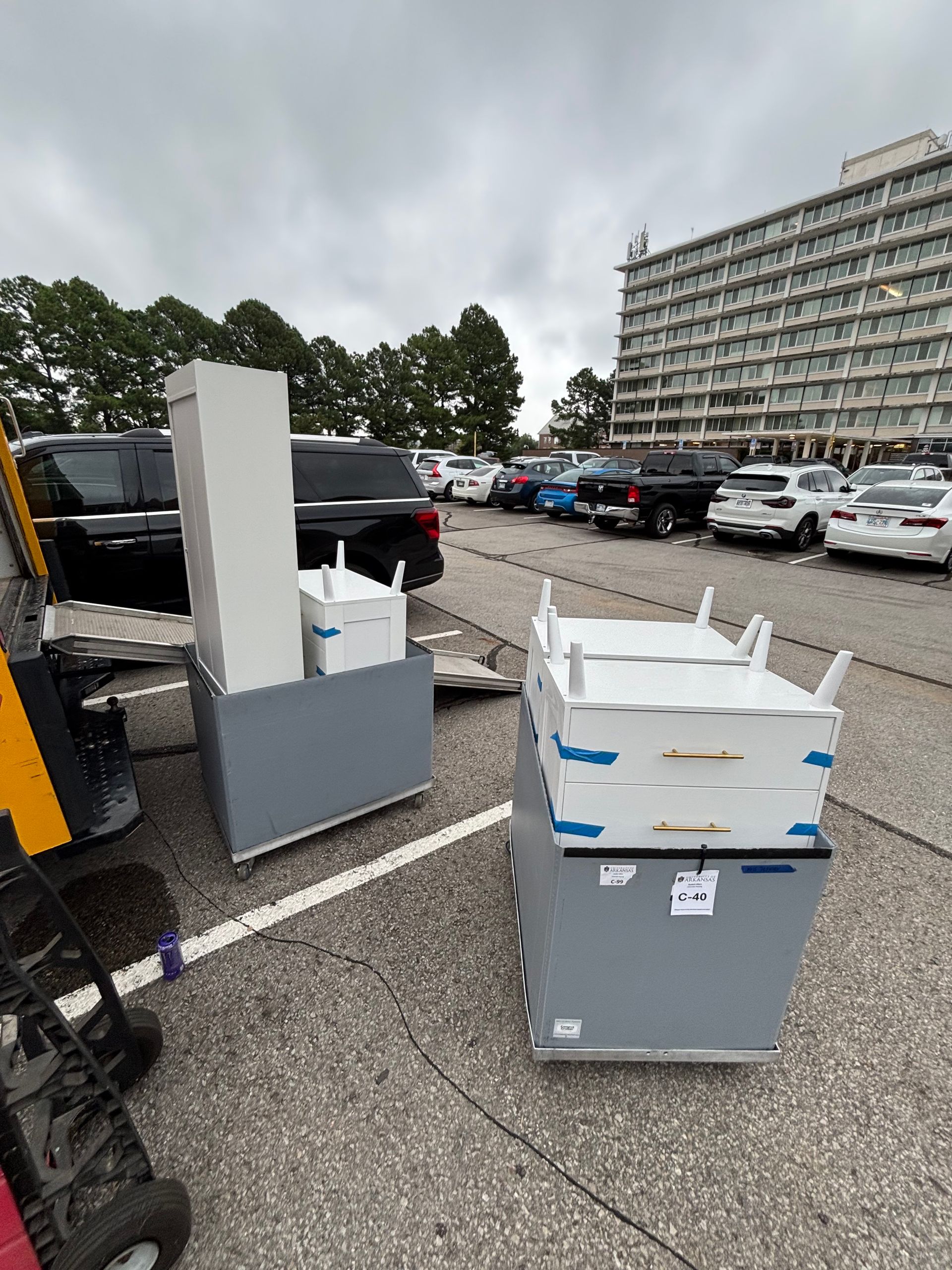 Two gray cabinets with white tops and components, parked on asphalt, near cars and a tall building.