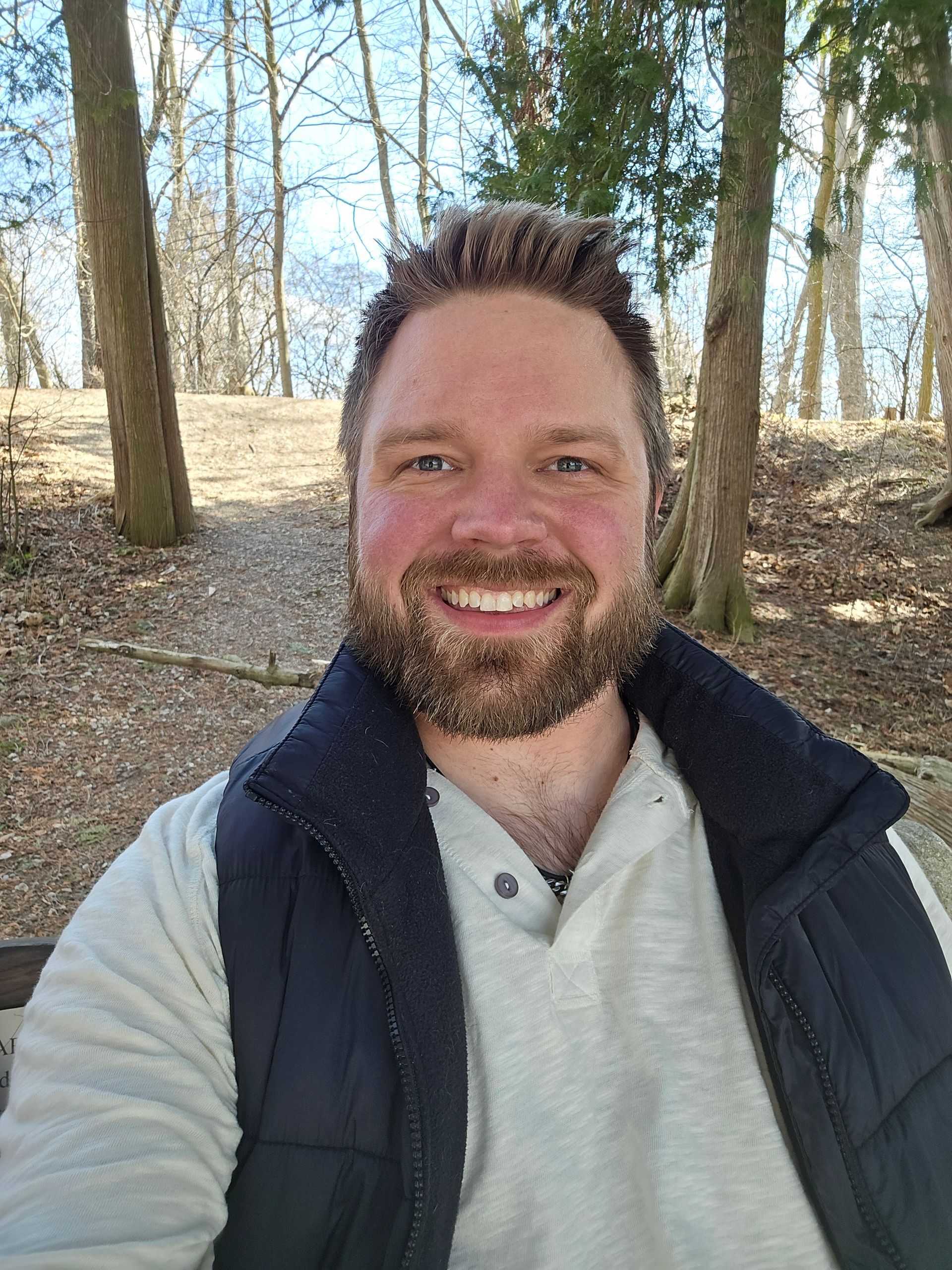 Man with a beard smiles outdoors, wearing a vest and light sweater, sitting on a bench; trees in background.