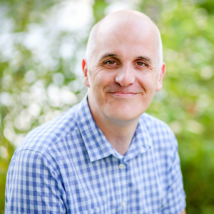 A bald man wearing a blue and white checkered shirt is smiling for the camera.