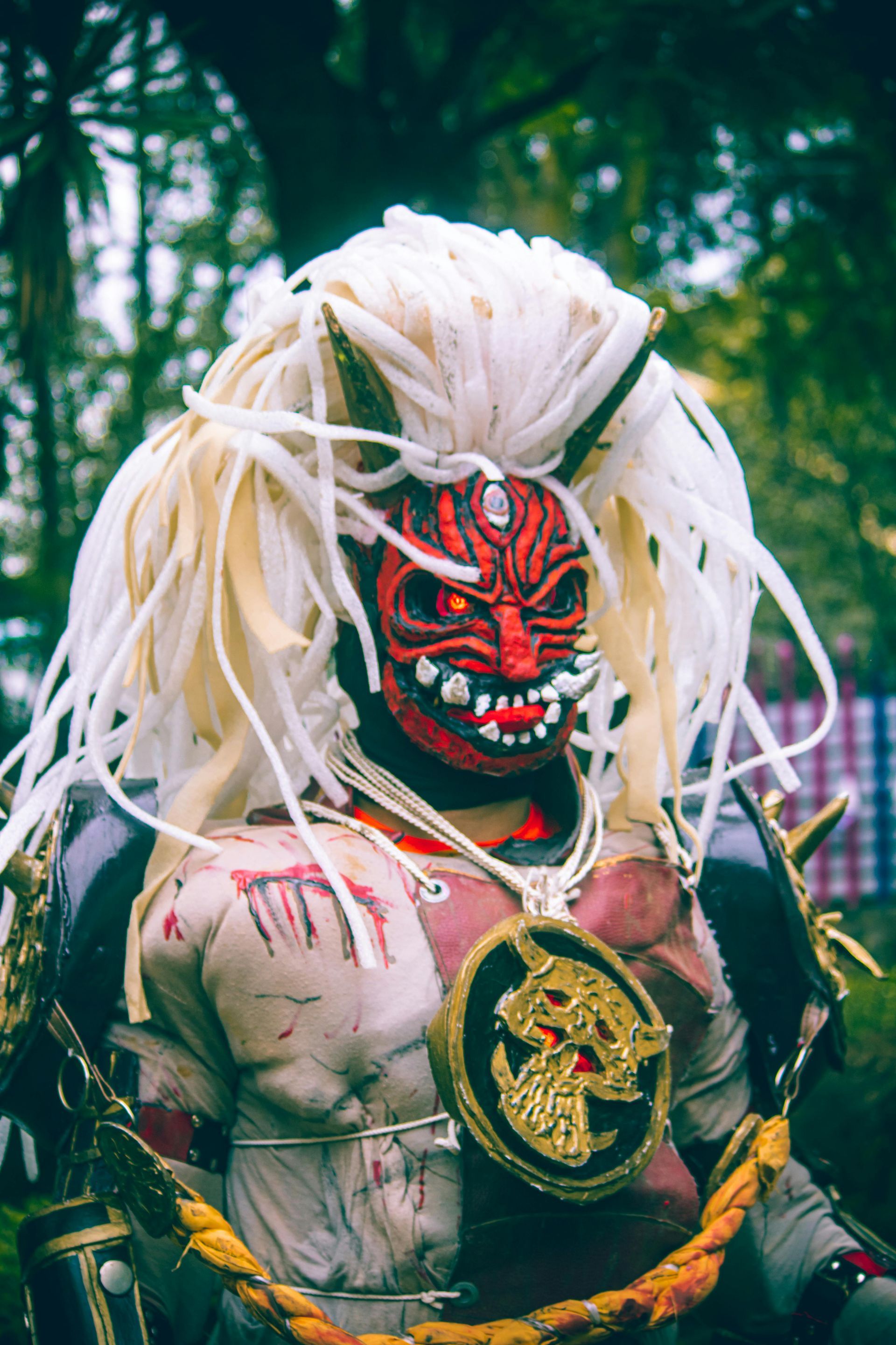 Person in a red demon mask with white hair, ornate necklace, and costume, set outdoors.