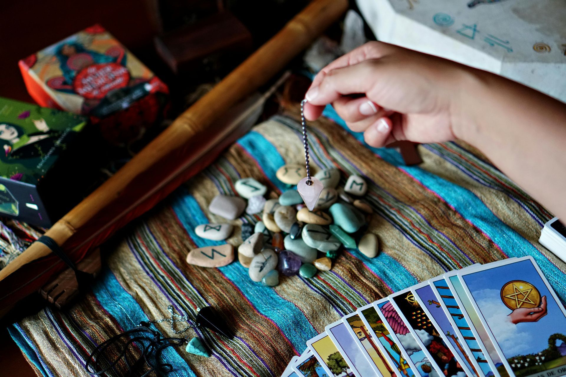 Person holds a heart-shaped pendulum above rune stones and tarot cards on a striped cloth, near a flute and boxes.