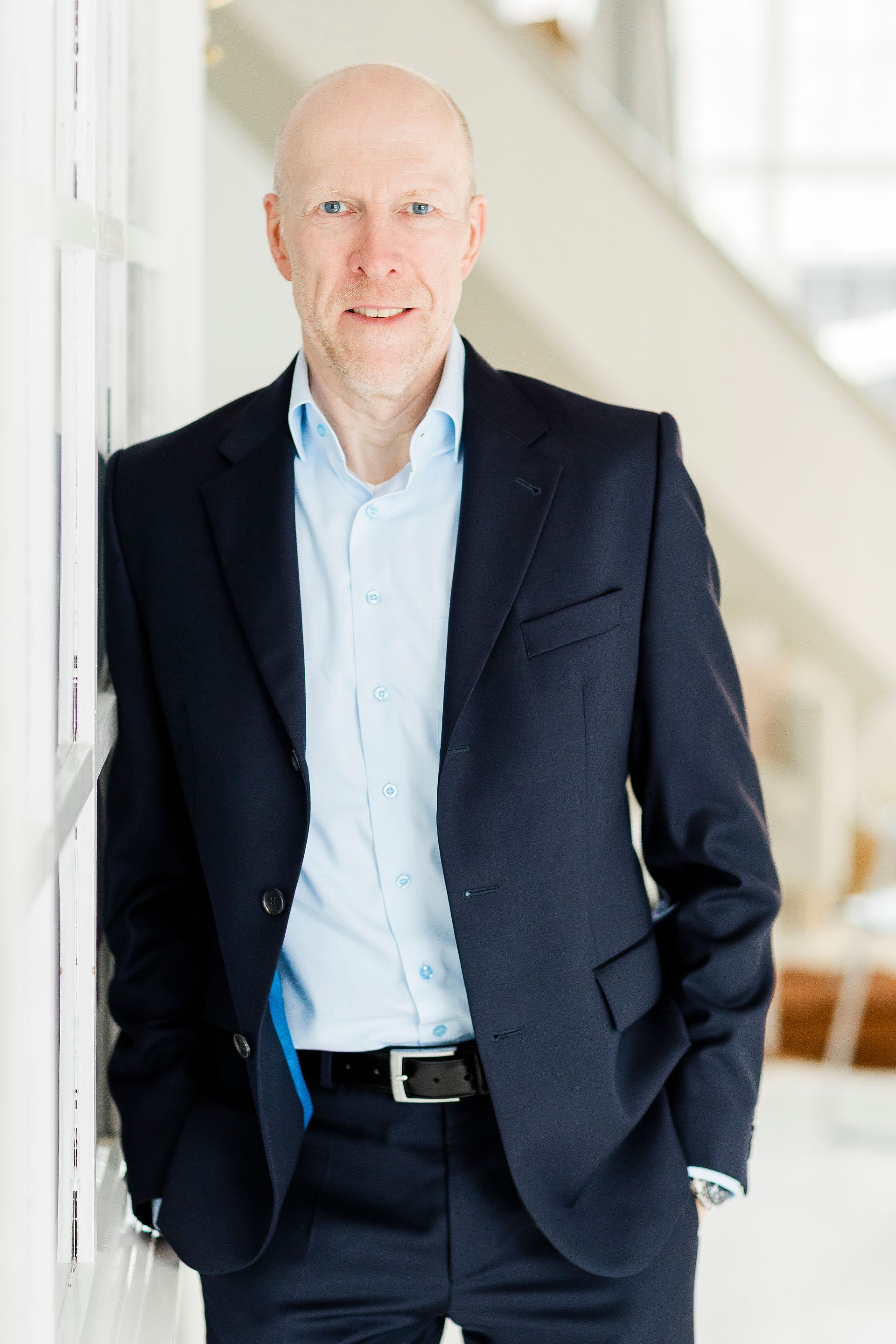 Ein Mann in einem dunkelblauen Blazer und einem hellblauen Hemd lehnt in einem hellen Büro an einer weißen Säule.