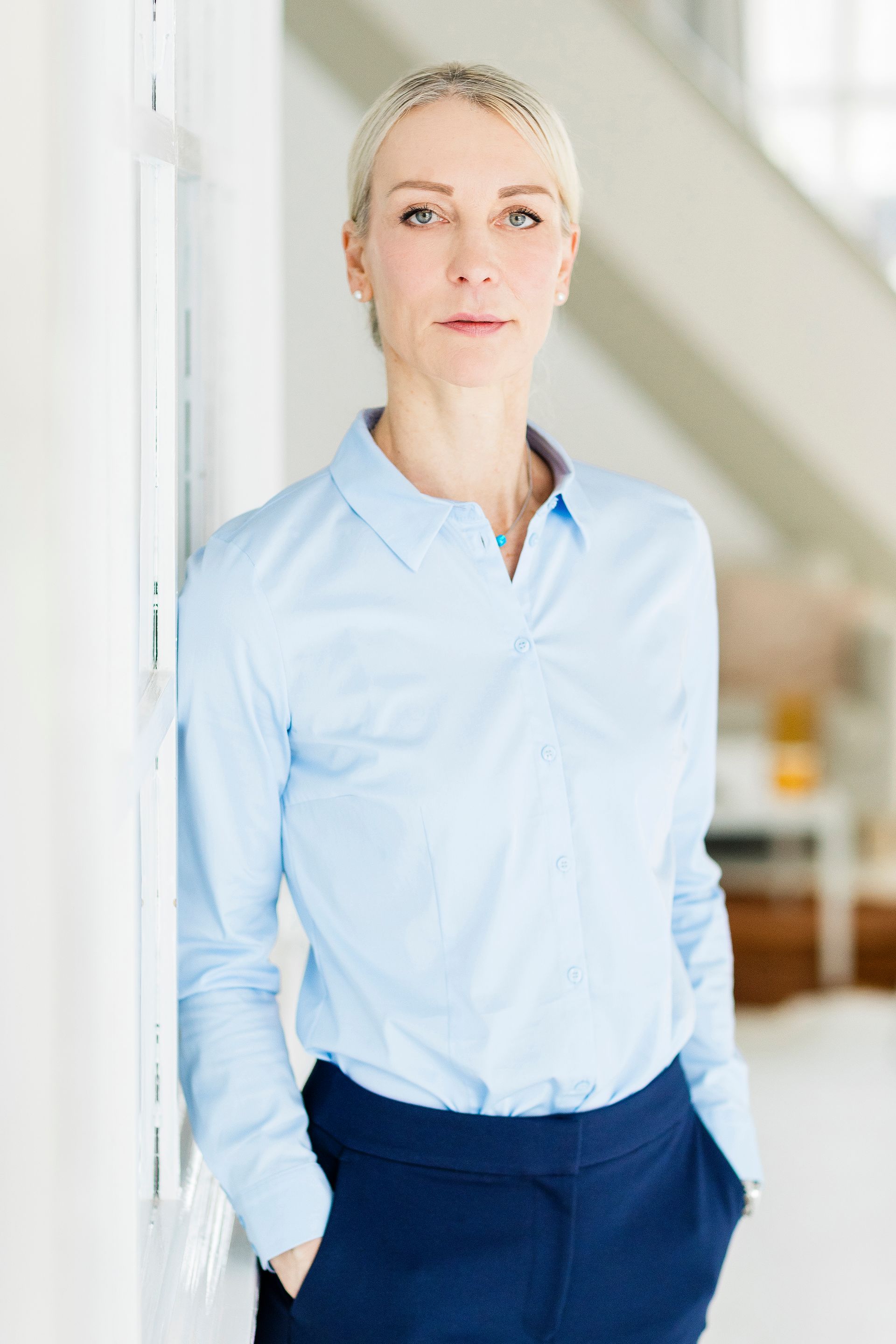 Eine Person mit kurzen blonden Haaren, einem hellblauen Hemd und einer dunkelblauen Hose lehnt an einer weißen Wand.