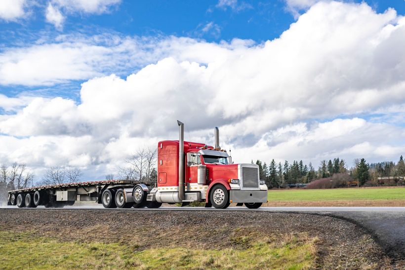 A red semi truck is driving down a road next to a field.