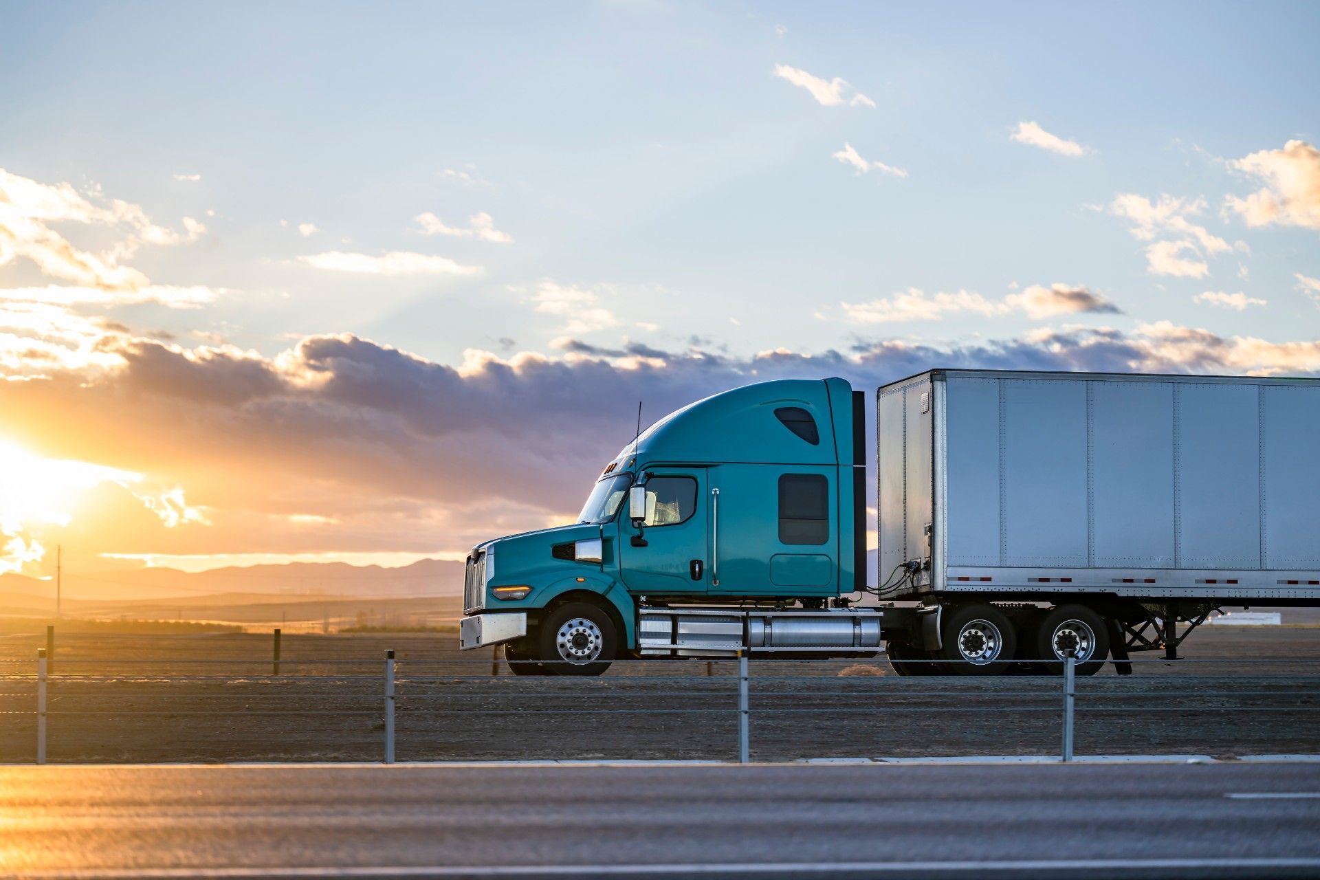 A blue semi truck is driving down a highway at sunset.