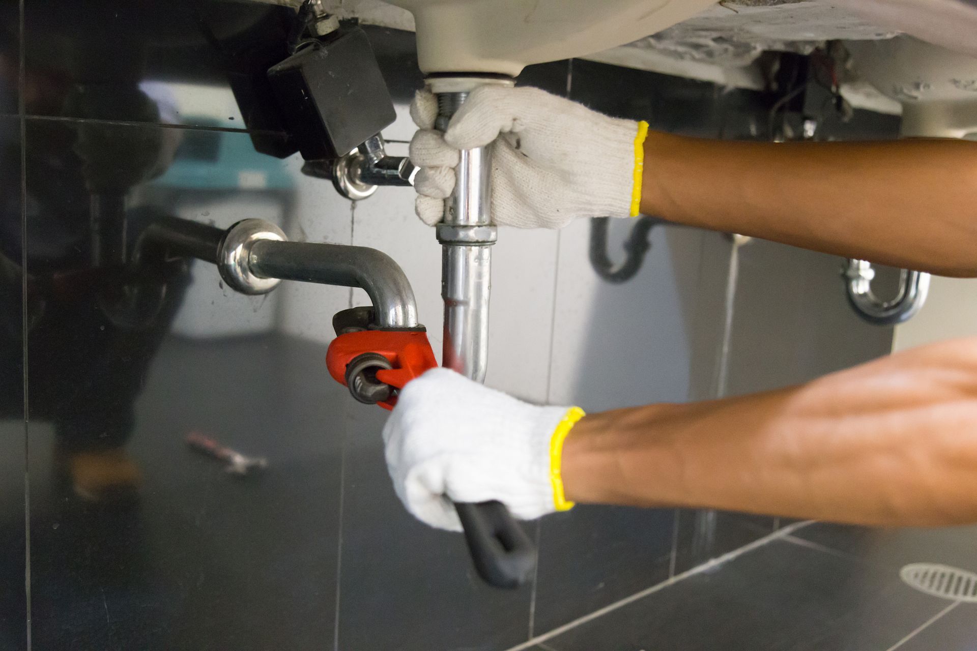 A person wearing white gloves uses a pipe wrench to tighten plumbing hardware under a sink.