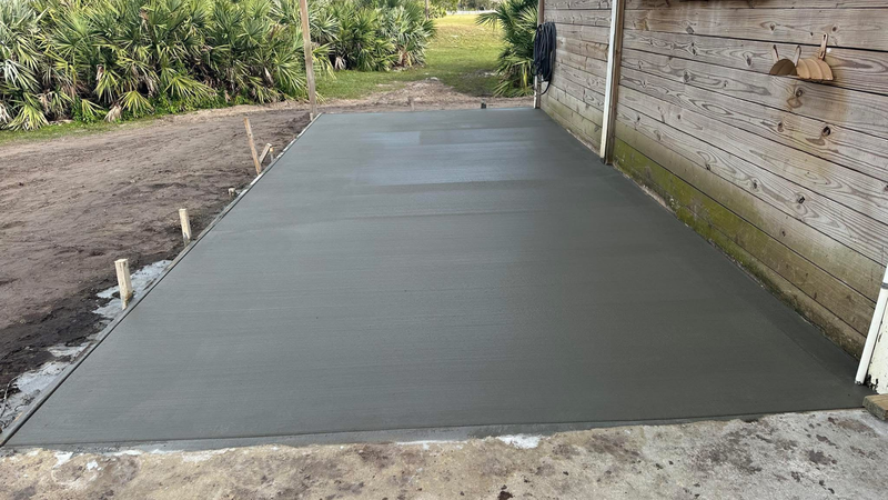 Freshly poured concrete patio next to a wooden building, surrounded by dirt and vegetation.