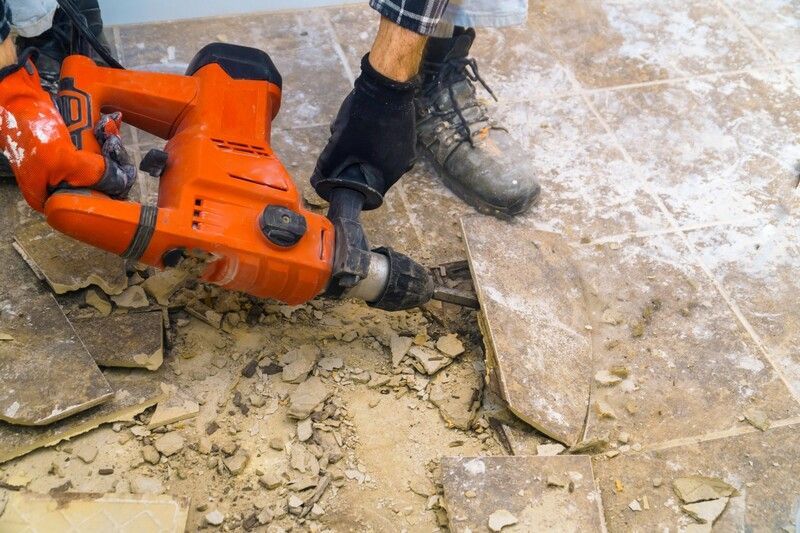 Person using an orange jackhammer to remove tile from a floor; debris is scattered around the work area.