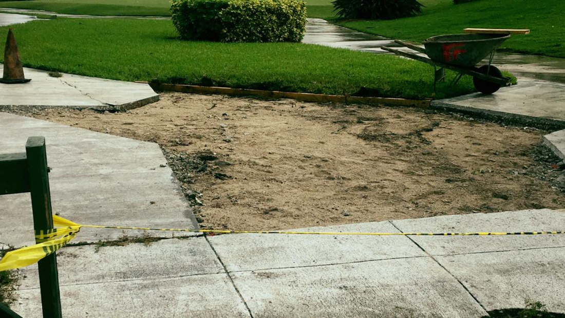 Path repair in progress: exposed dirt patch surrounded by concrete sidewalk, safety tape, wheelbarrow in background.