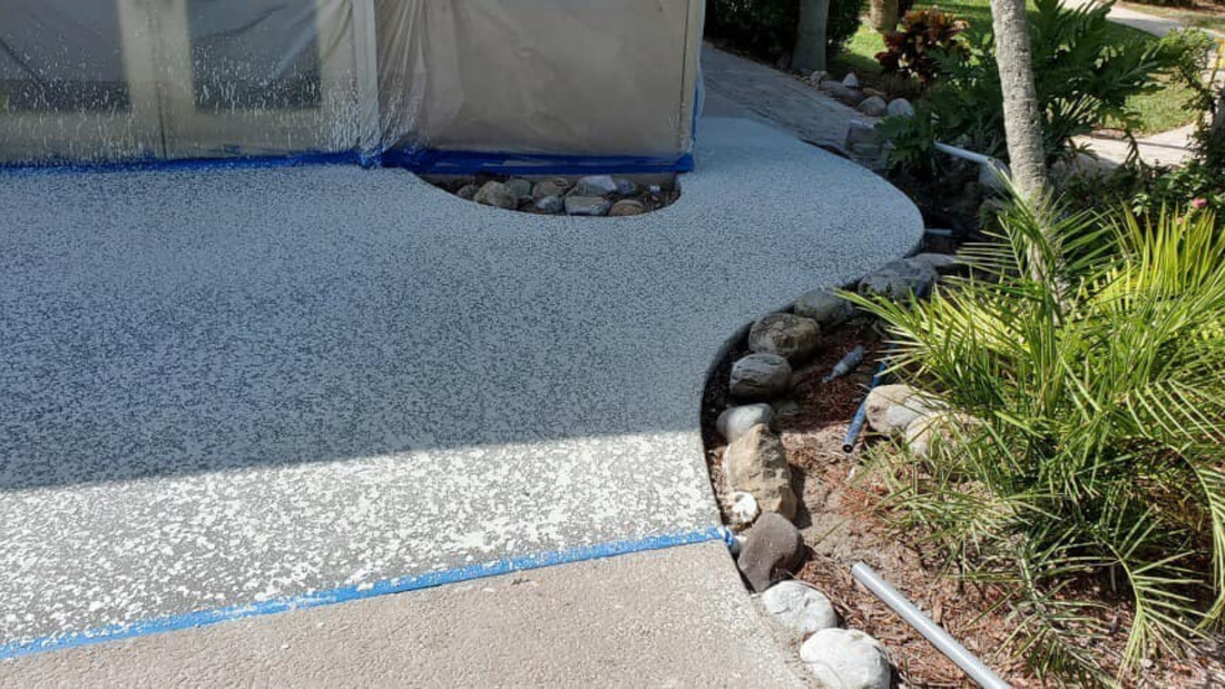 Light-colored, textured concrete patio edging a small garden with rocks and plants, adjacent to a house with blue tape.