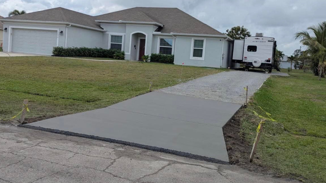 A light blue house with a gray cement driveway. An RV is parked in the gravel portion.