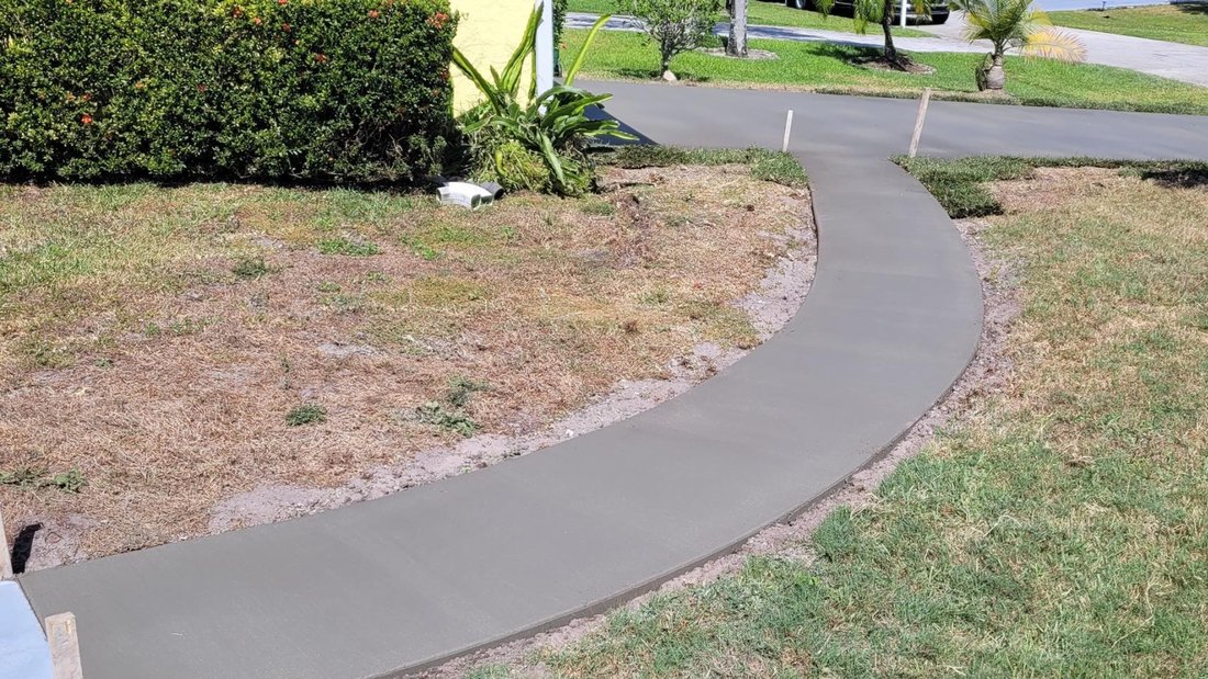A newly poured concrete walkway curves through a grassy yard, with a hedge and street in the background.