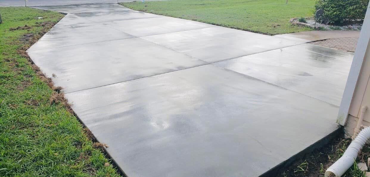 Newly poured concrete driveway with a wet, reflective surface, surrounded by green grass.