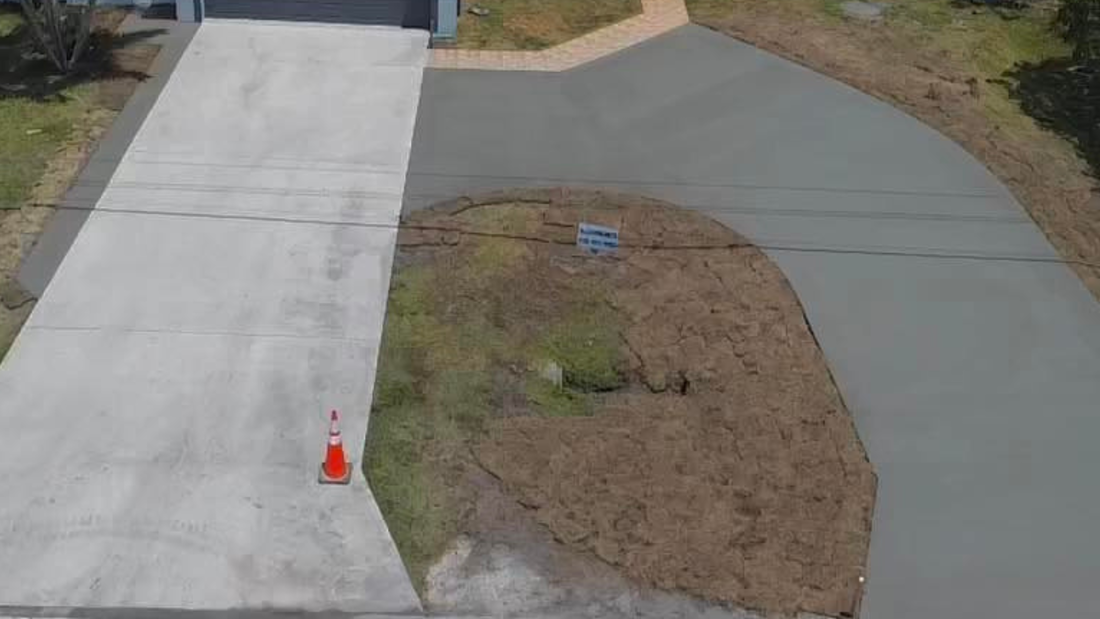 Driveway with concrete and asphalt sections, a traffic cone, and a grassy median.