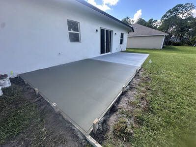 Newly poured concrete patio next to a white house, on a grassy lawn.