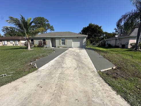 New concrete driveway leading to a gray house, with grass on either side under a blue sky.