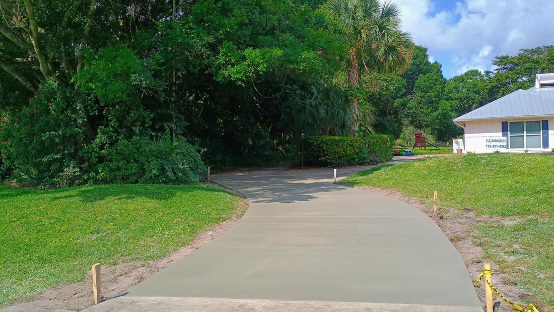 A freshly poured concrete pathway curves through a grassy area, leading toward a building and dense green trees.