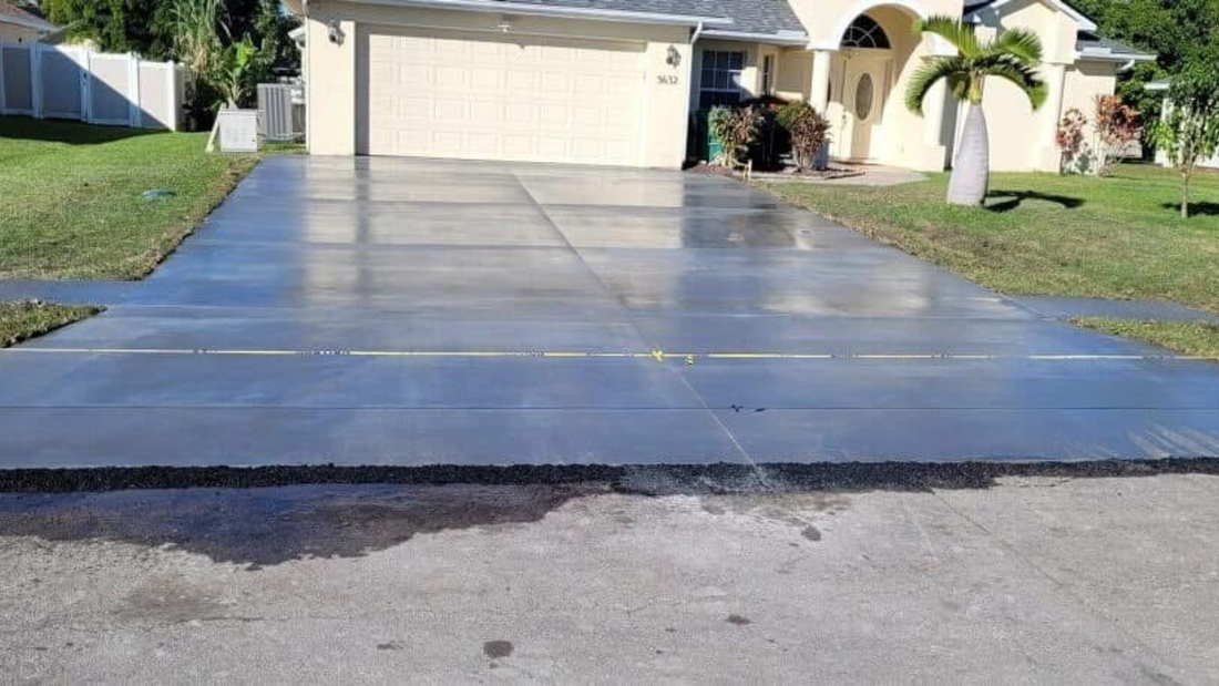 Newly sealed, wet, concrete driveway in front of a light-colored house with green lawn and a palm tree.