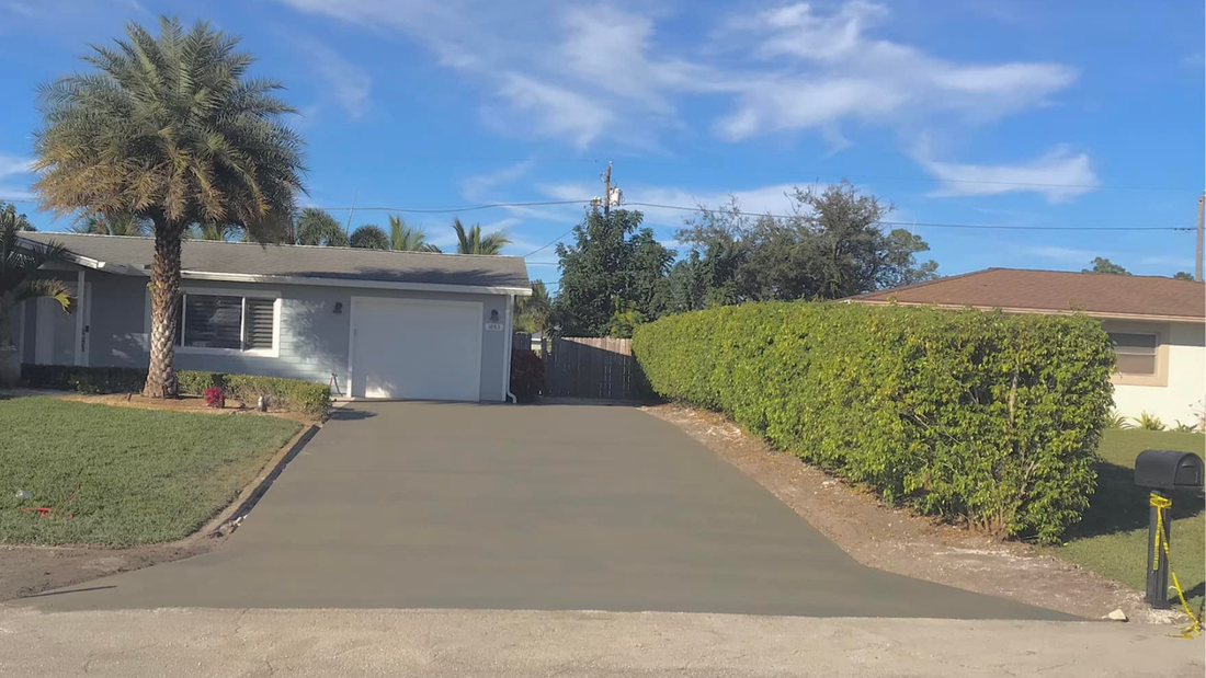 Driveway of a light blue house, palm tree, green hedge, and clear sky.