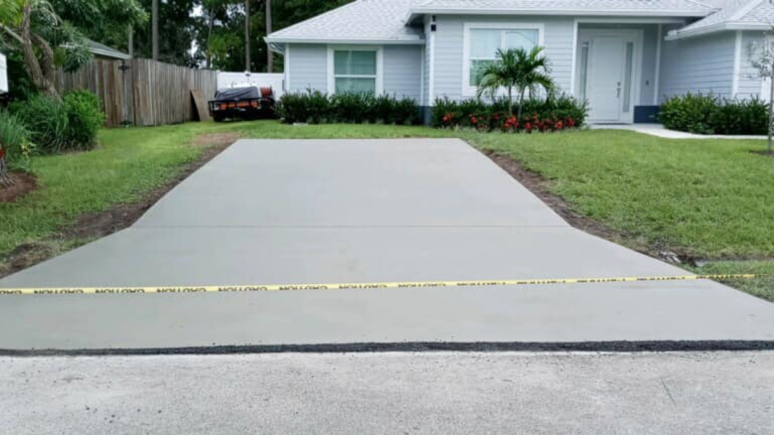 Newly poured concrete driveway in front of a light blue house, with caution tape and green grass.