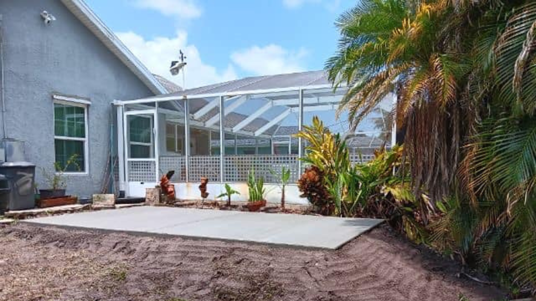 Backyard patio with a screened enclosure. Gray house, concrete patio, plants, and blue sky.