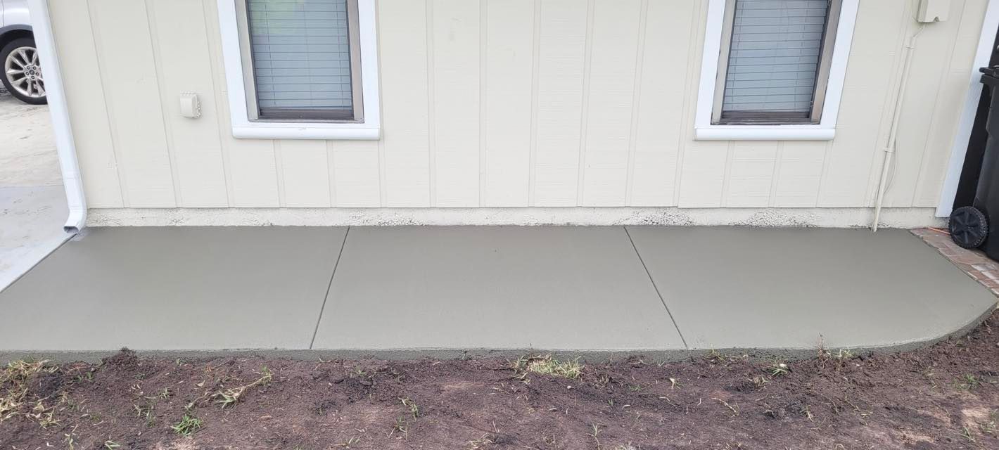 Newly poured concrete patio next to a house with two windows and surrounding soil.