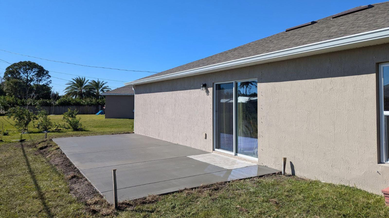Newly poured concrete patio next to a house with sliding glass doors, surrounded by grass under a blue sky.