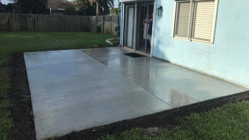 Newly poured concrete patio next to a turquoise house, with grass and a sliding glass door.