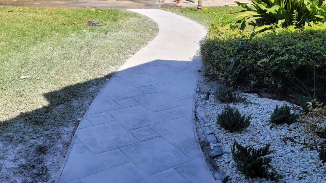 Curving concrete pathway through a grassy yard and beside a hedge with white stones.