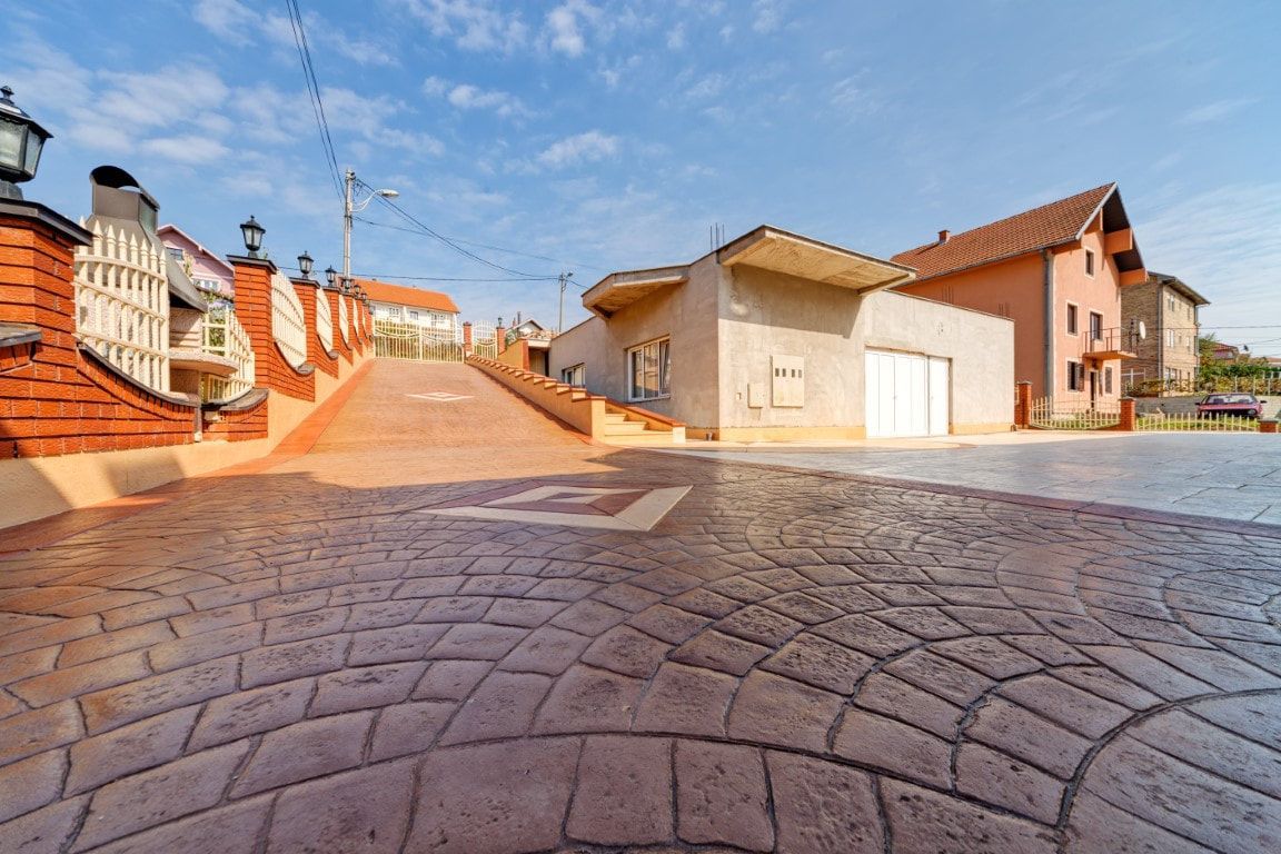 Brick-patterned concrete driveway leading uphill to buildings with red tile roofs under a blue sky.