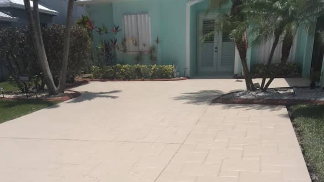 A beige driveway leads to a turquoise house with white trim and double doors; palm trees and shrubbery on the sides.