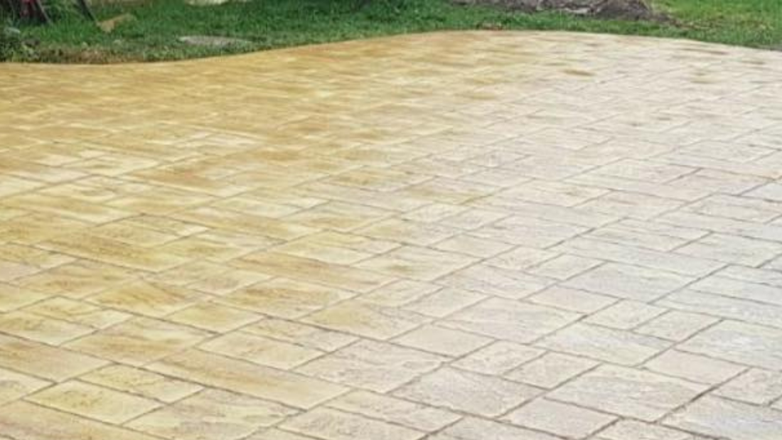 Concrete patio, tan and light brown, with brick-like pattern, next to green grass.