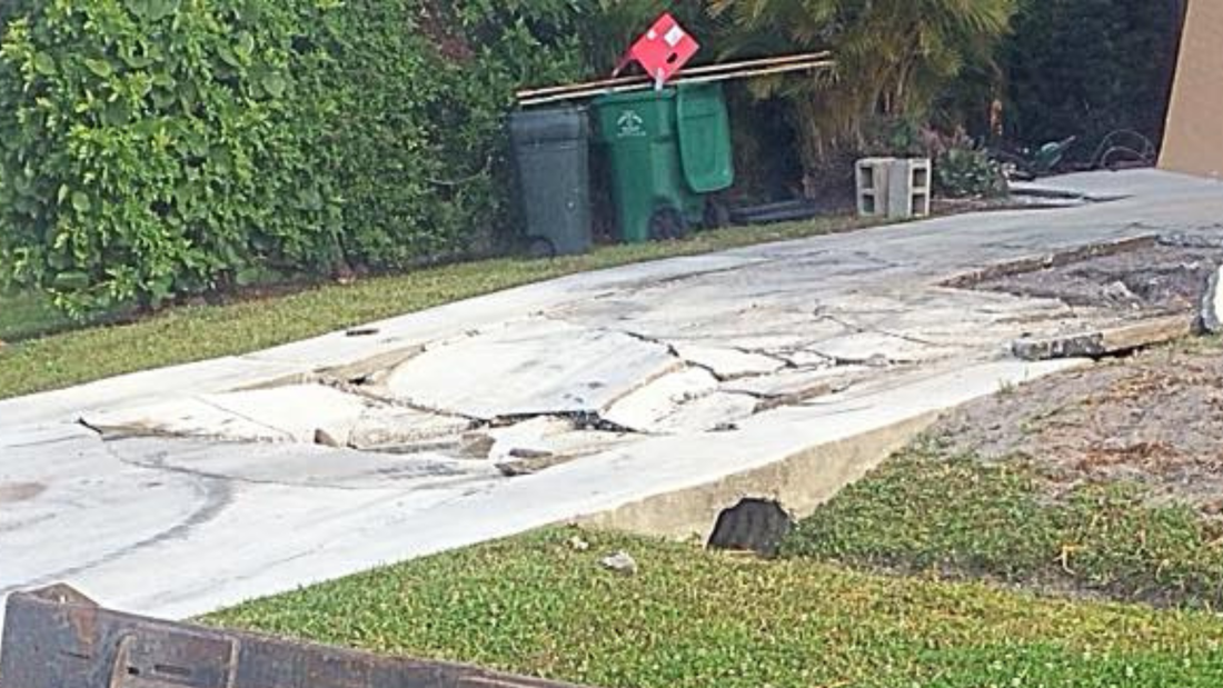 Damaged concrete driveway with large cracks, flanked by grass and trash cans.